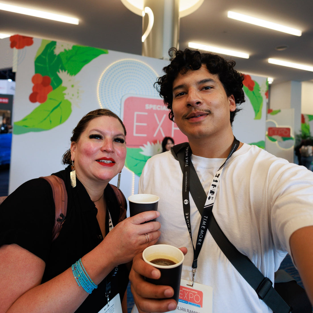 Julian Ramirez and Luci Ramirez of Humboldt Bay Coffee smiling and holding cups of coffee at the Specialty Coffee Association Expo in Houston, with the expo sign and floral decor in the background.
